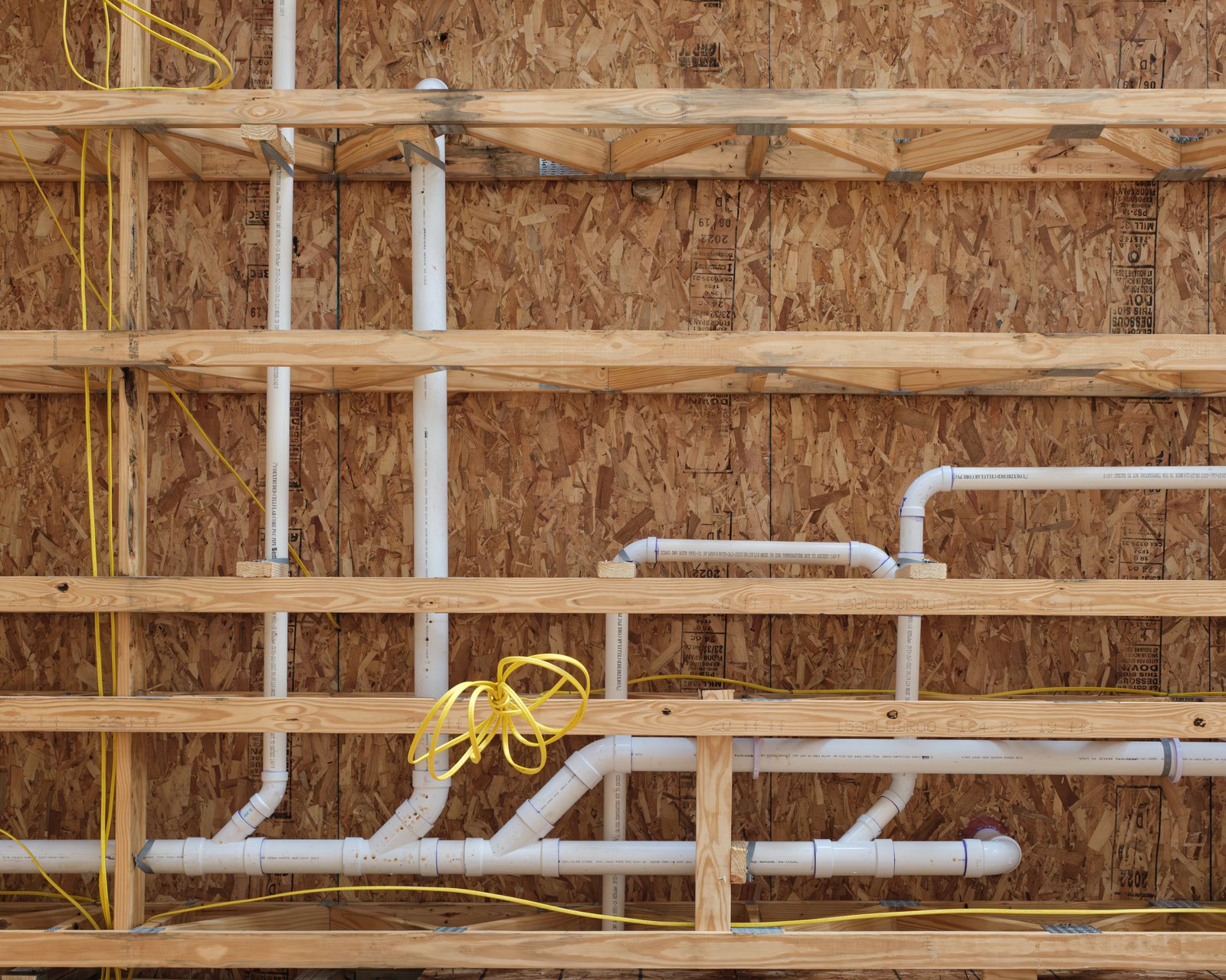 Pre-drywall inspection view of an unfinished new construction interior showing exposed wood framing, white PVC plumbing rough-in, and yellow electrical wiring against OSB sheathing.
