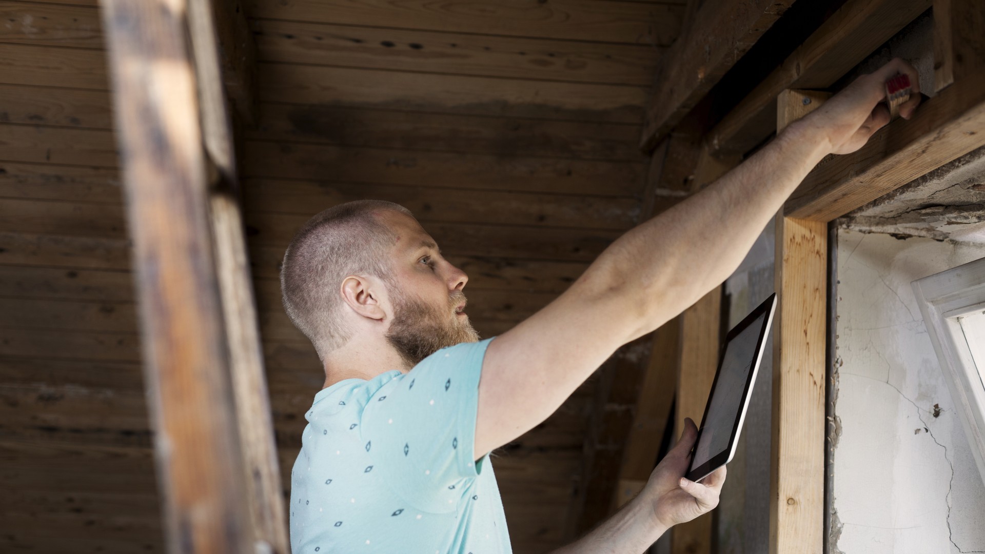 A home inspector examining an older home's wood framing and window area and documenting the condition on a tablet