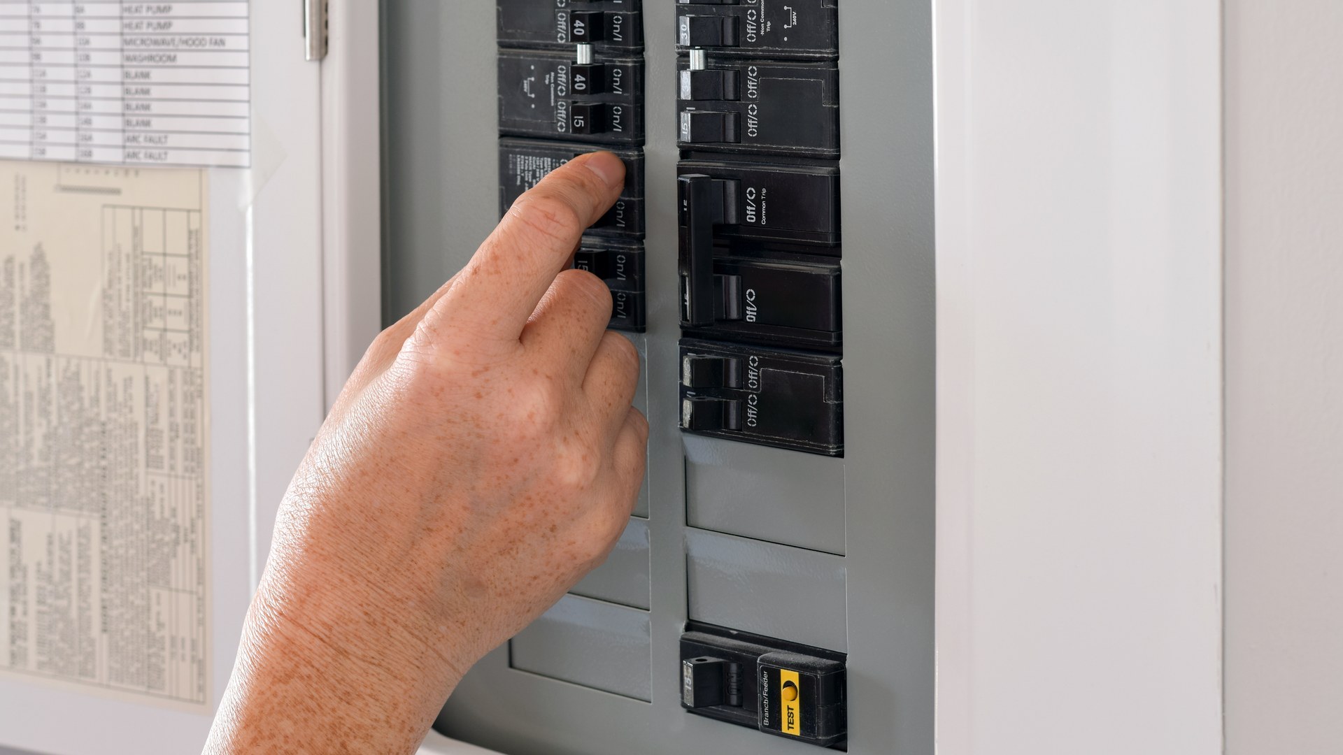 A hand at an open residential electrical panel with the circuit directory listing heat pump and spare blank slots.