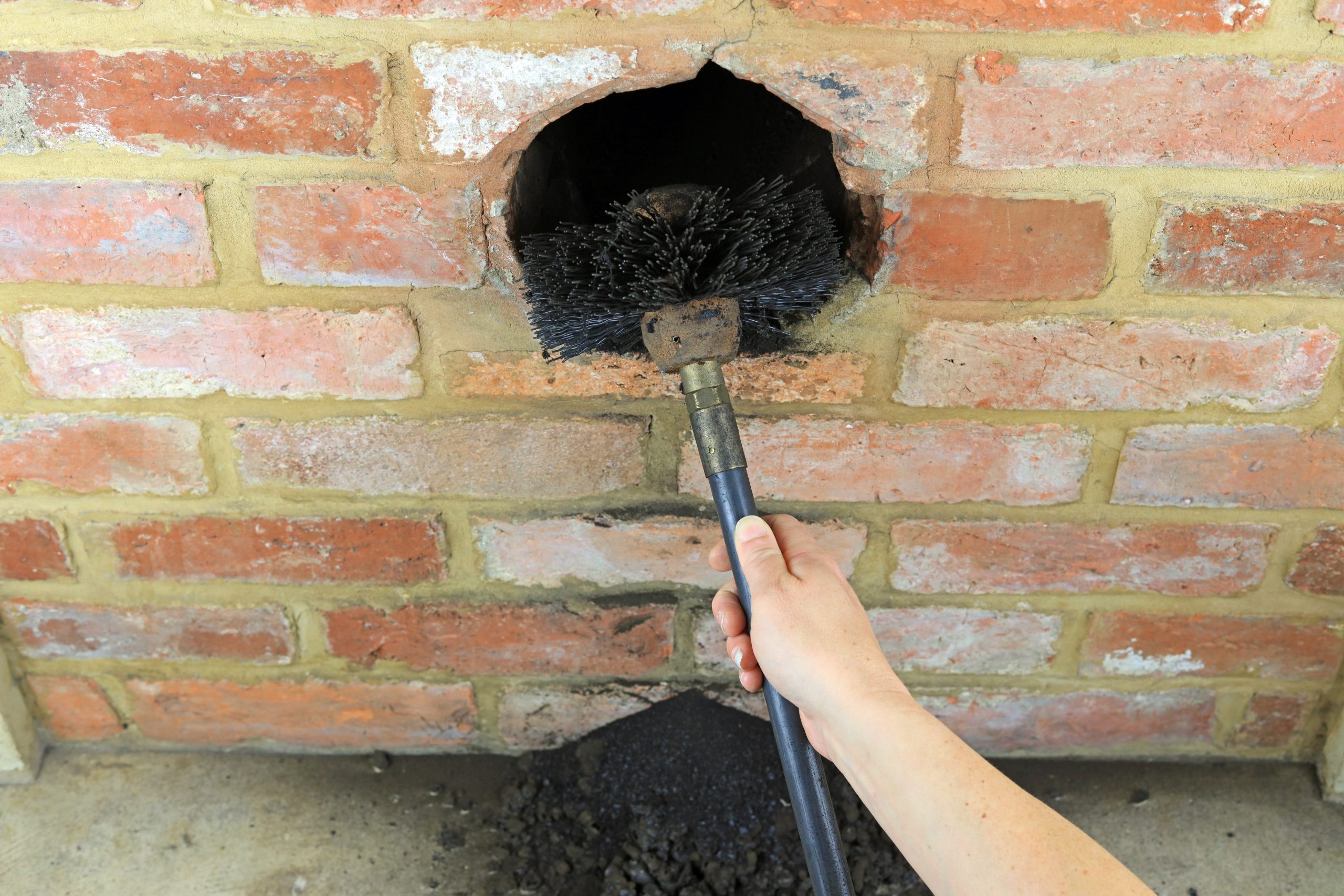 Bristle brush on a rod being pushed up into the flue opening of an old brick chimney with soot on the hearth below