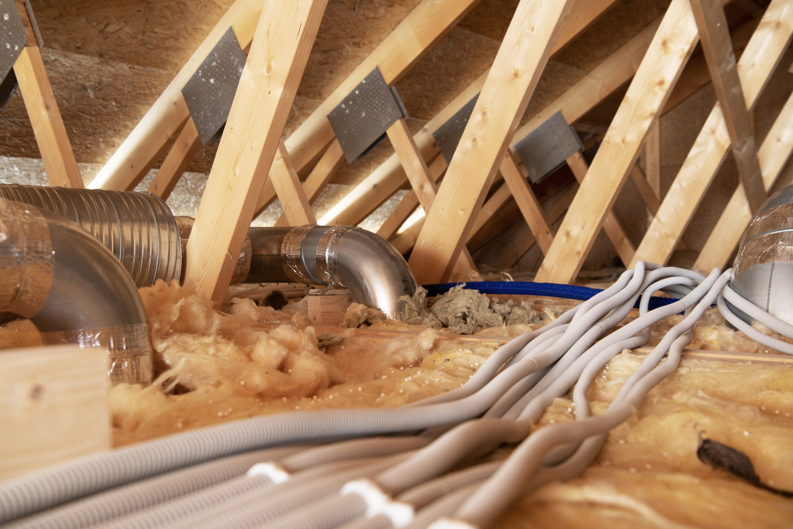 Residential attic interior showing roof framing, batt insulation over the ceiling joists and metal duct elbows running across it