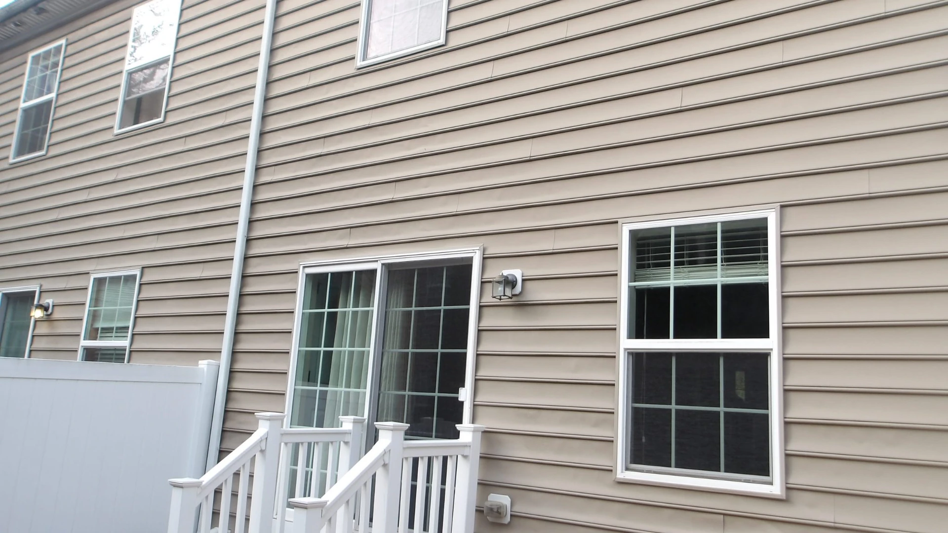 Rear of an attached multi-unit building showing one unit's sliding door and stair landing beside a neighboring unit behind a privacy fence