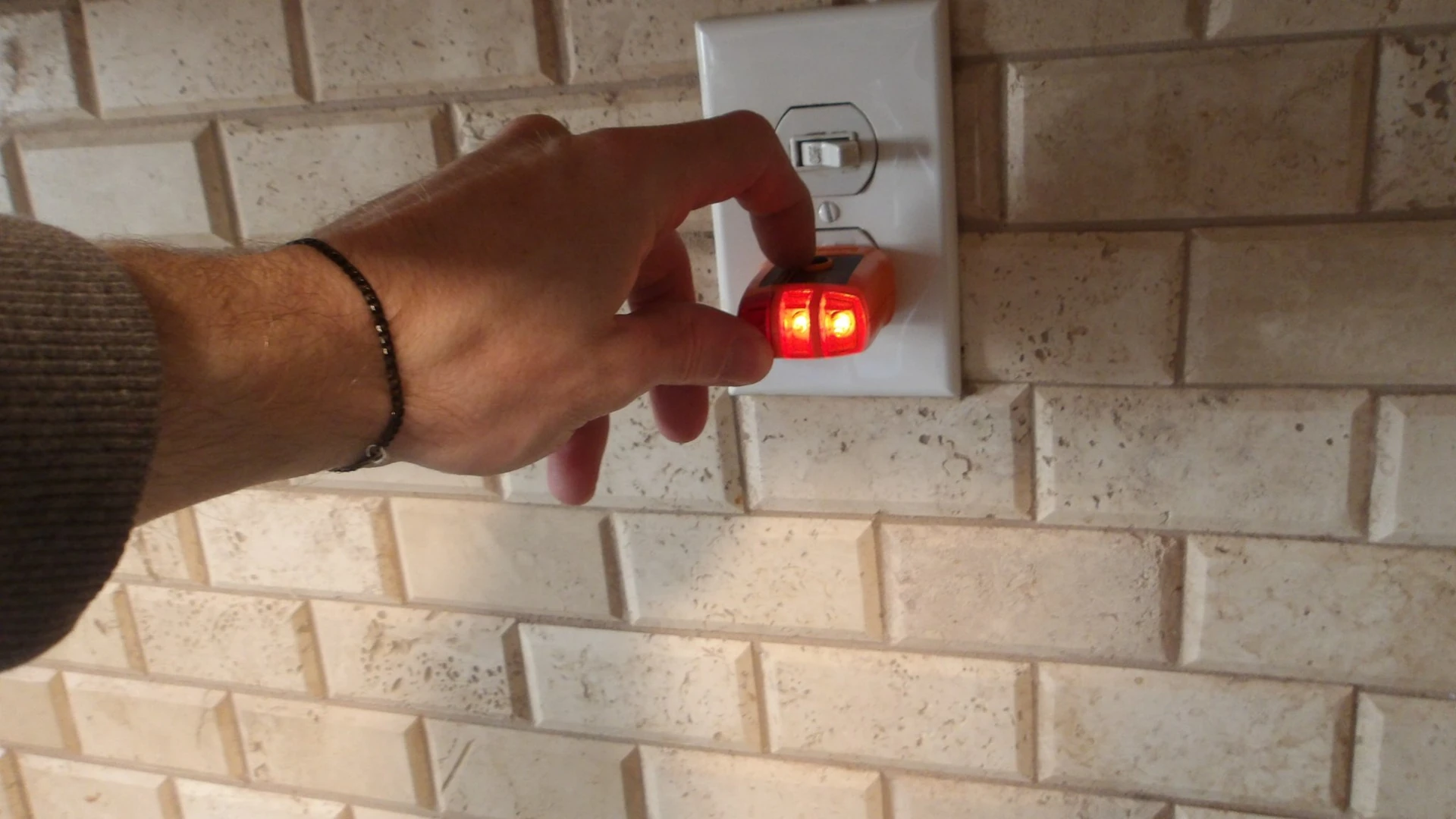 An inspector's hand plugs a lit outlet tester into a receptacle on a tiled backsplash.
