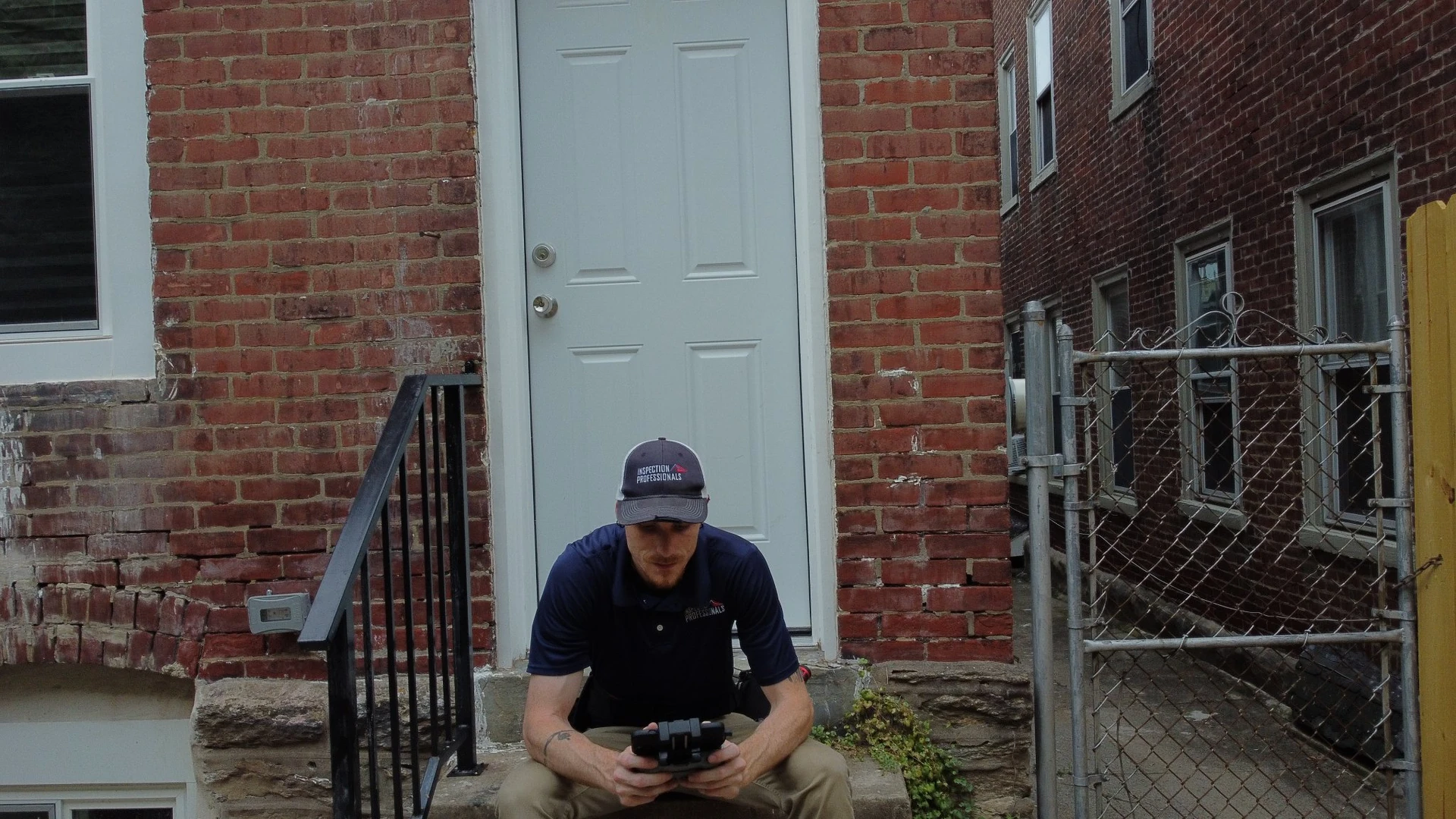 An Inspection Professionals inspector sits on the stone steps of an older brick home, looking down at a drone controller.
