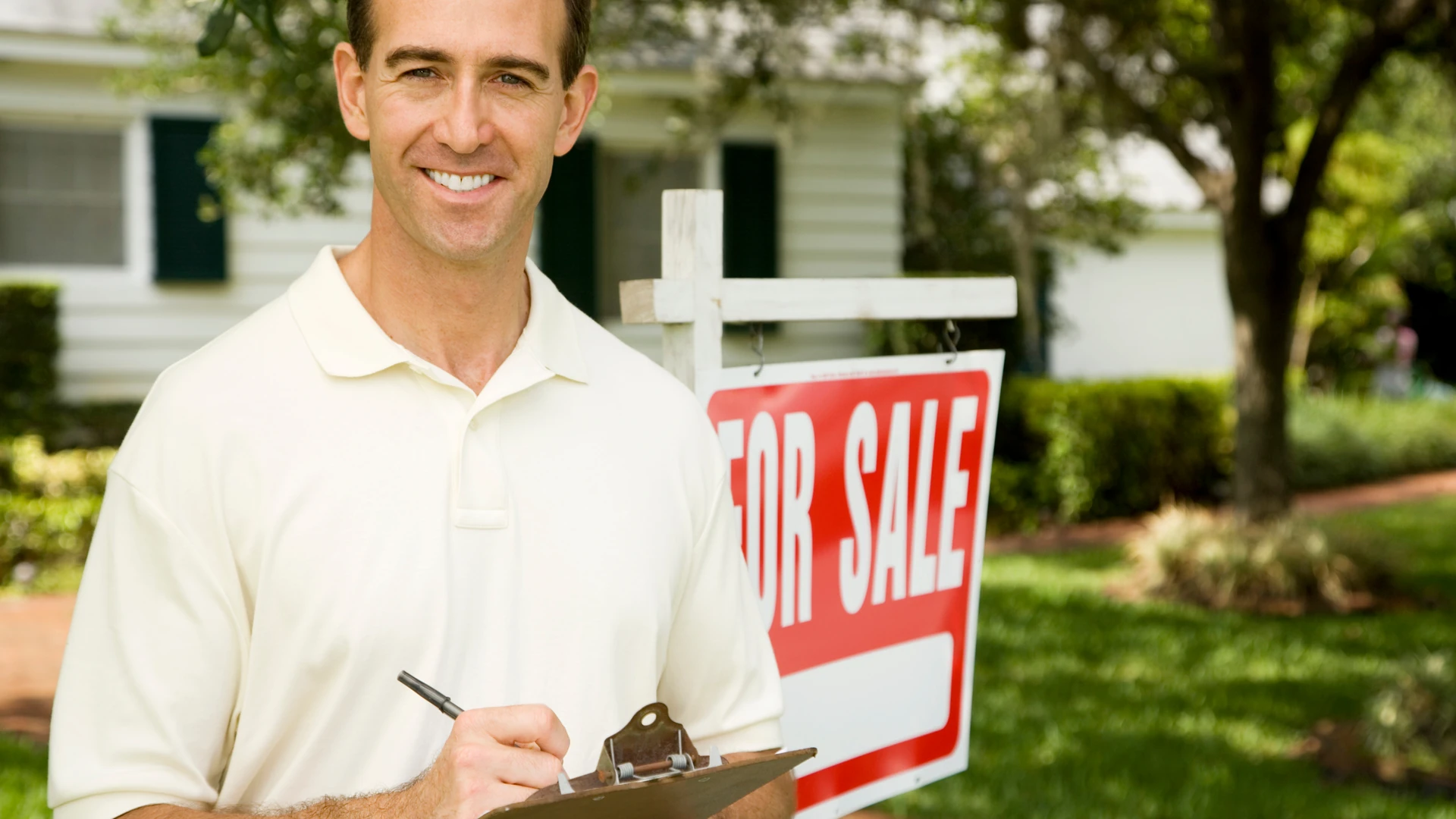 Smiling man taking notes on a clipboard beside a For Sale sign on the lawn of a white house