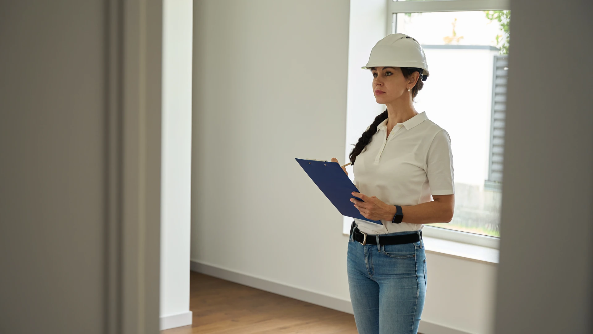 Inspector in a white hard hat taking notes on a blue clipboard while checking an empty room in a home