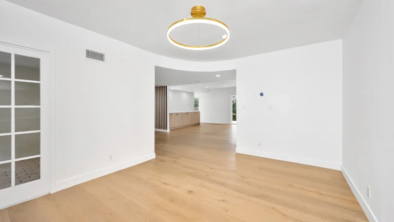 Empty room with white walls and light wood plank flooring, a round gold ceiling light, a glass-paned door at the left and a doorway opening to a room with wood cabinets in the background
