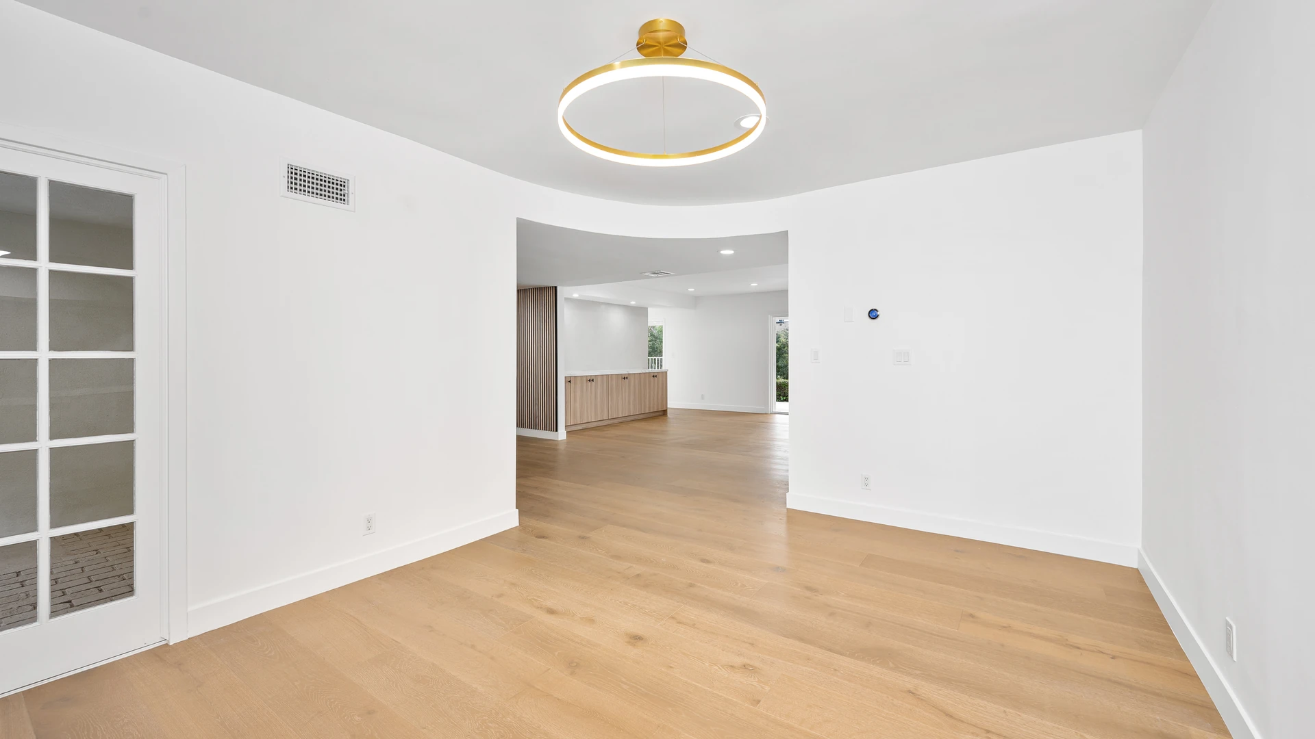 Empty room with white walls and light wood plank flooring, a round gold ceiling light, a glass-paned door at the left and a doorway opening to a room with wood cabinets in the background