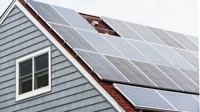 Gable end of a house with gray siding and a white-framed window beside a large array of solar panels mounted on the red roof