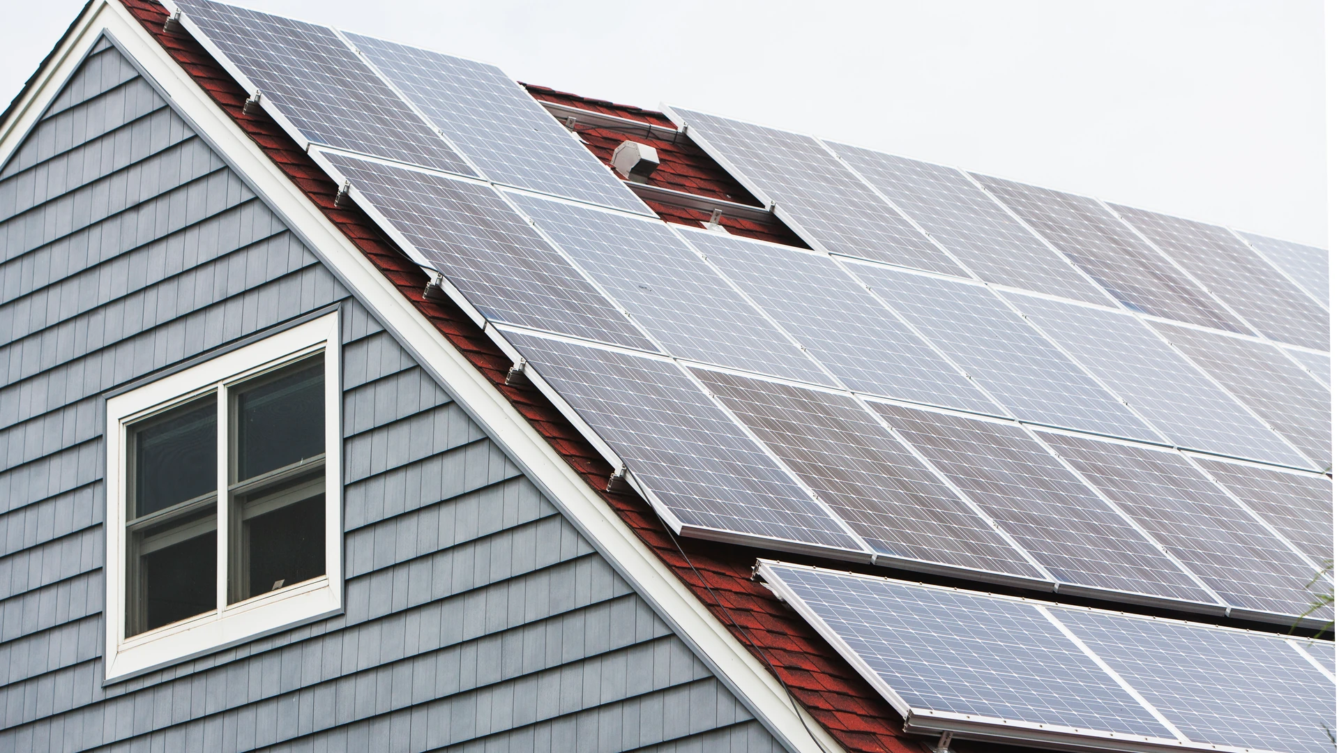 Gable end of a house with gray siding and a white-framed window beside a large array of solar panels mounted on the red roof