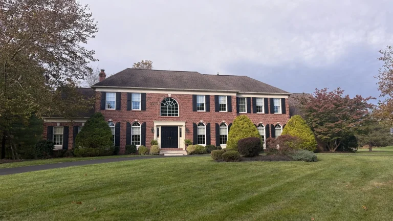 Two-story red-brick house with black shutters, an arched window above the front door, trimmed shrubs and a wide lawn under a cloudy sky