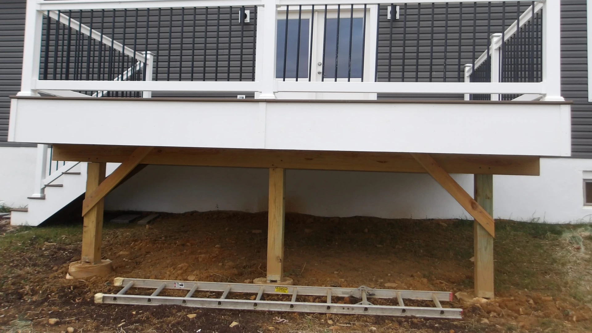 Elevated deck with white railings, wooden support posts with diagonal bracing standing on bare soil, and a ladder lying on the ground beneath it