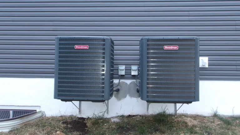Two gray outdoor air-conditioning units with red logo labels sit on brackets against gray horizontal siding above a white foundation wall, with a small white fixture on the siding at the right and a window-well grate at the lower left