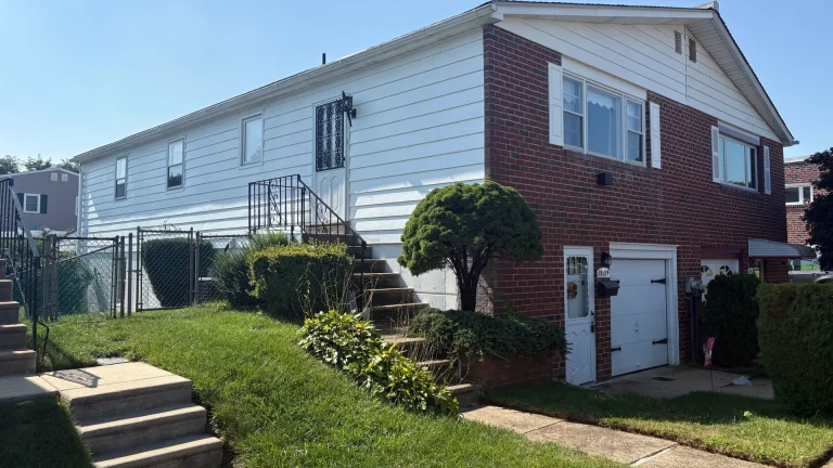 Side of a house with a brick lower level with a white garage door and white siding above, and a lawn, shrubs, a railed stair and concrete steps in front