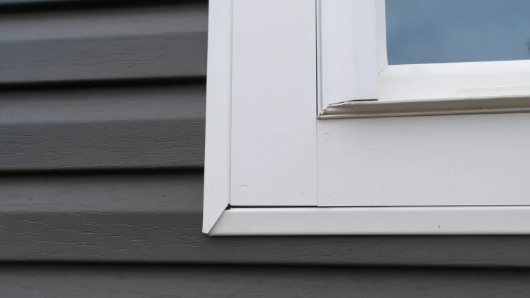 Lower left corner of a white-framed window with white trim around it, set against gray horizontal siding, with a blue-gray reflection in the glass at the top right