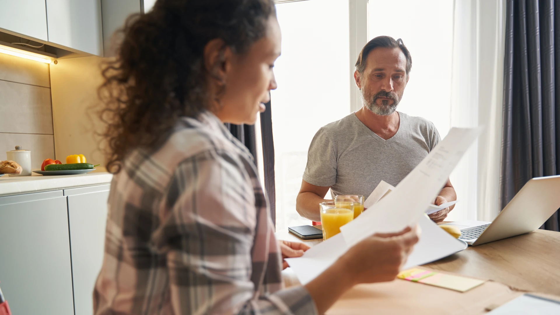 A woman and a man at a kitchen table, each holding printed pages, with a laptop and two glasses of orange juice on the table