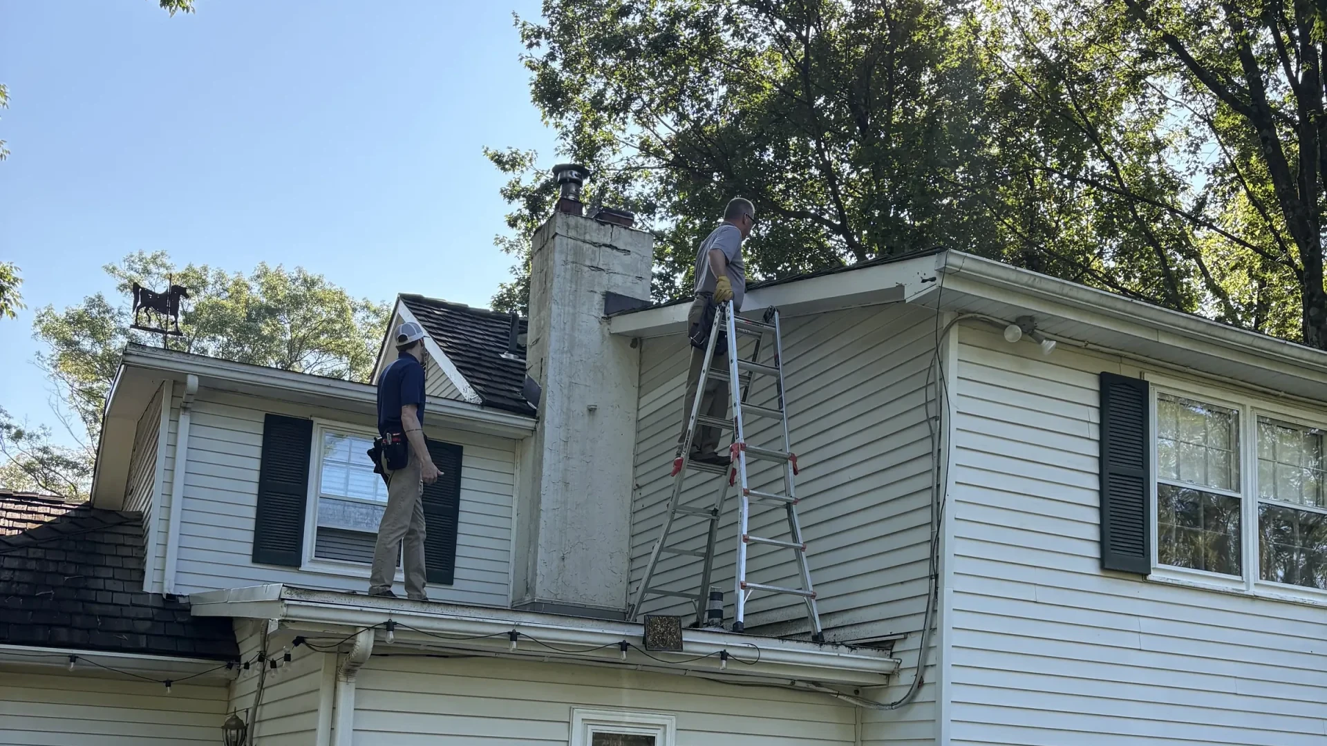 Two people at the roof edges of a white-sided house with a tall chimney: one in a navy shirt and white cap stands at the lower roof edge looking up, and another stands on an aluminum ladder leaned against the upper wall at the eave, with gutters along both roof edges and a horse weather vane at the left