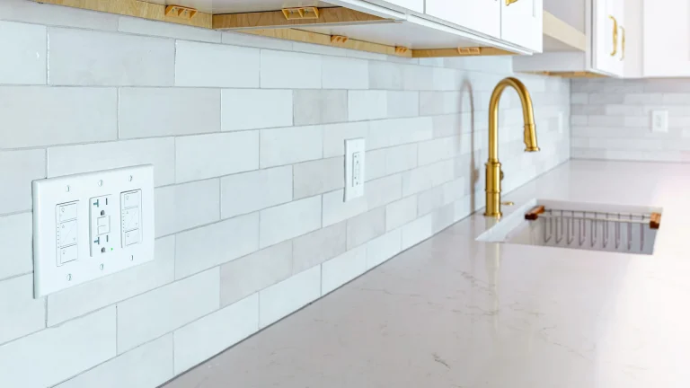 White wall plate with two dimmer switches and a GFCI outlet on a tile kitchen backsplash beside a gold faucet