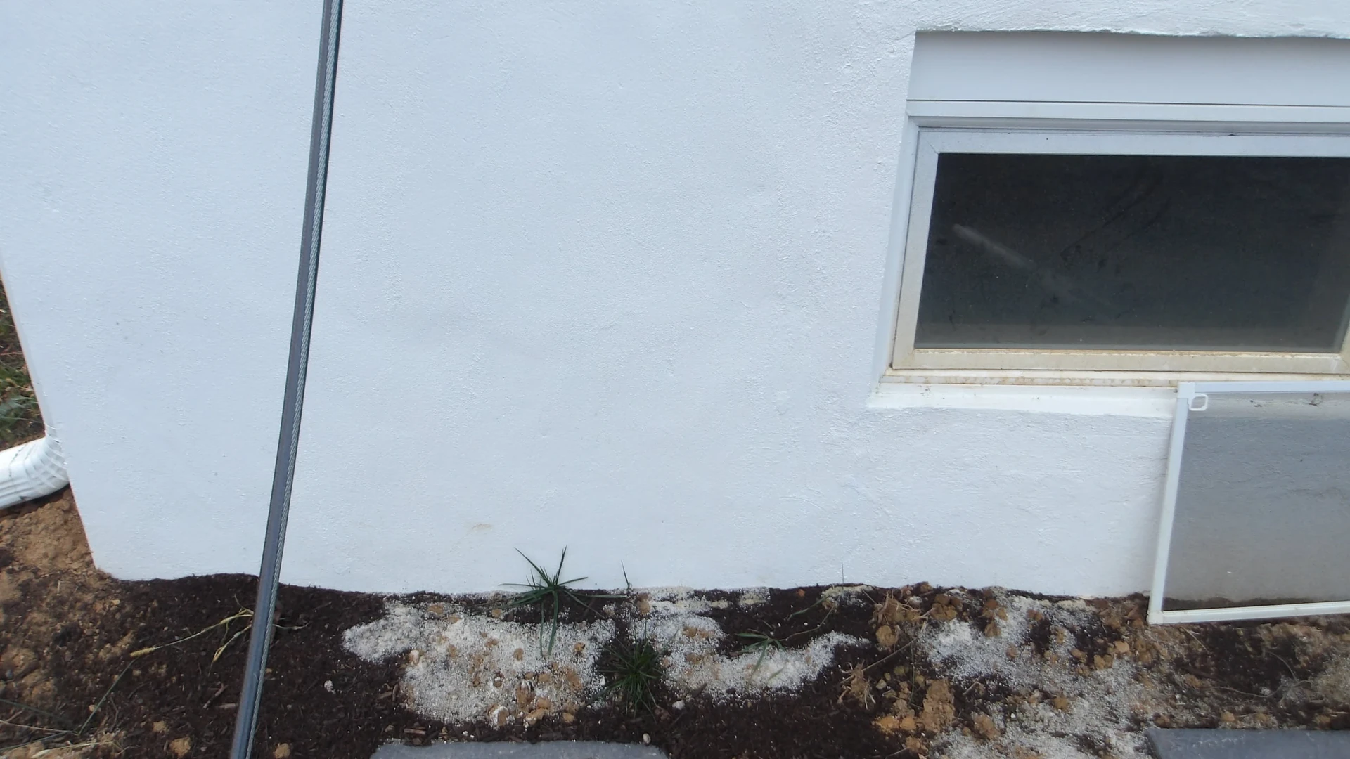 A white textured foundation wall with a small low window at upper right and a window screen leaning against the wall beneath it, a thin dark pole at left, and dark soil with white granules along the base
