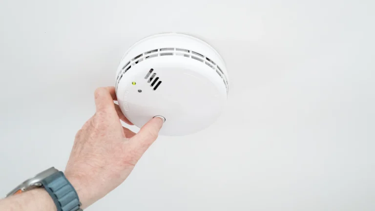 A hand with a gray-blue wristwatch band reaches up to press the button on a round white ceiling-mounted smoke and carbon monoxide detector on a plain white ceiling