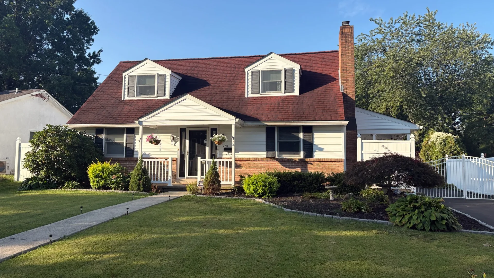 White house with a dark red shingle roof, two dormer windows, a brick chimney, a covered front porch and a landscaped front lawn with a concrete walkway